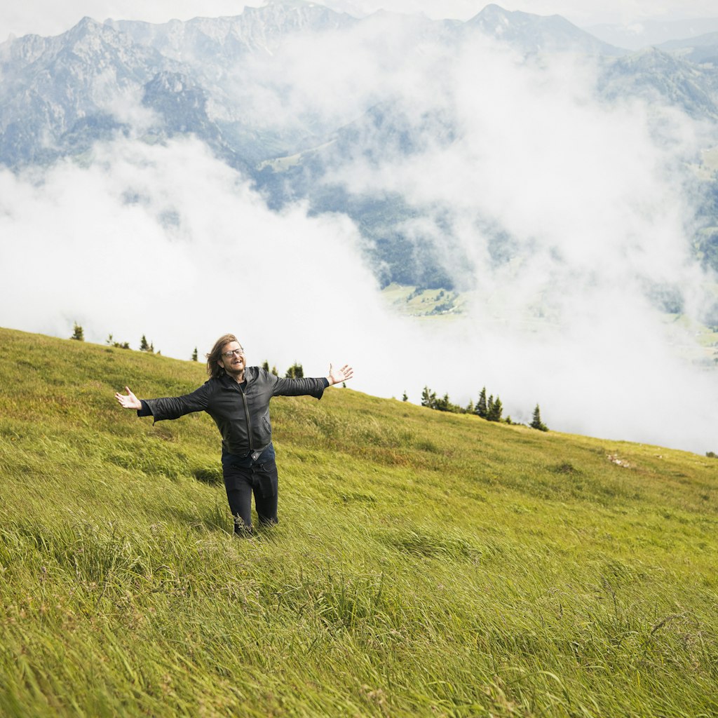 Man stands in a grassy meadow with his arms wide apart on the side of Schafberg Mountain.