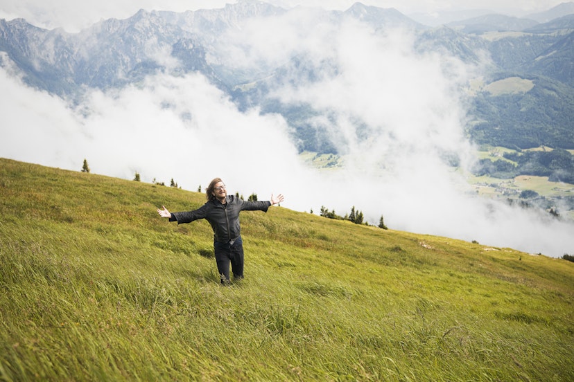 Man stands in a grassy meadow with his arms wide apart on the side of Schafberg Mountain.