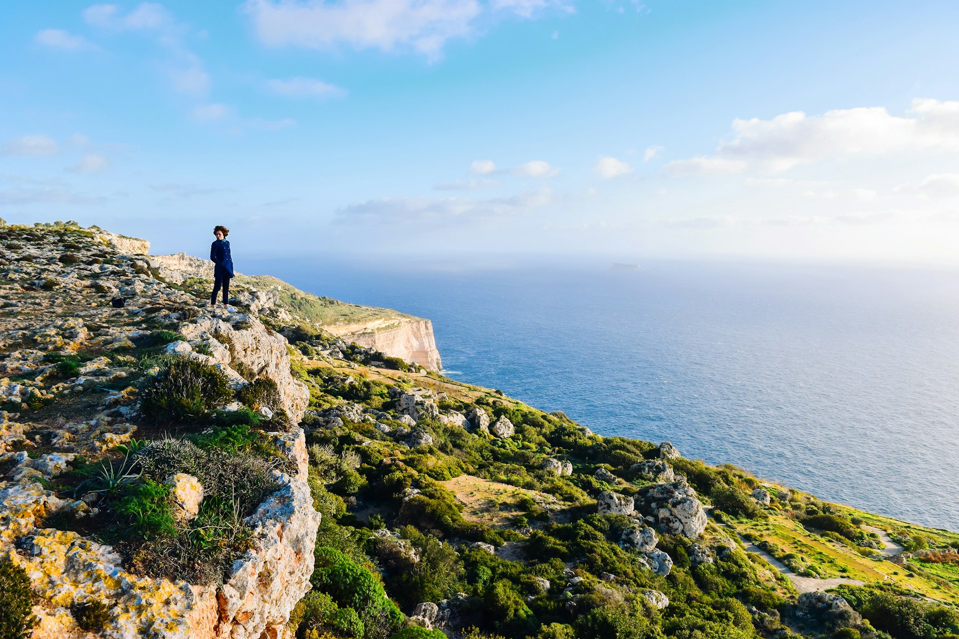 A young woman stands at a height overlooking the Dingli cliffs and the Mediterranean Sea. Her hair is blown by the wind.