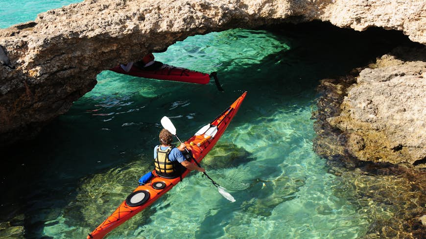 Two kayakers in turquoise water navigate their way under a natural limestone arch, Comino Islands, Malta.
