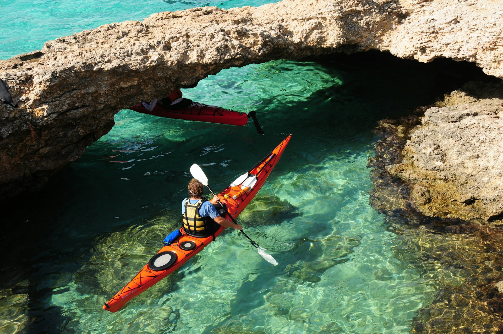 Two kayakers in turquoise water navigate their way under a natural limestone arch, Comino Islands, Malta.