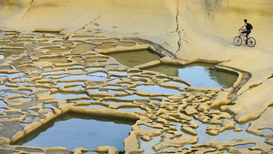 A solo cyclist pedals round a flat yellowish landscape in Gozo's salt pans