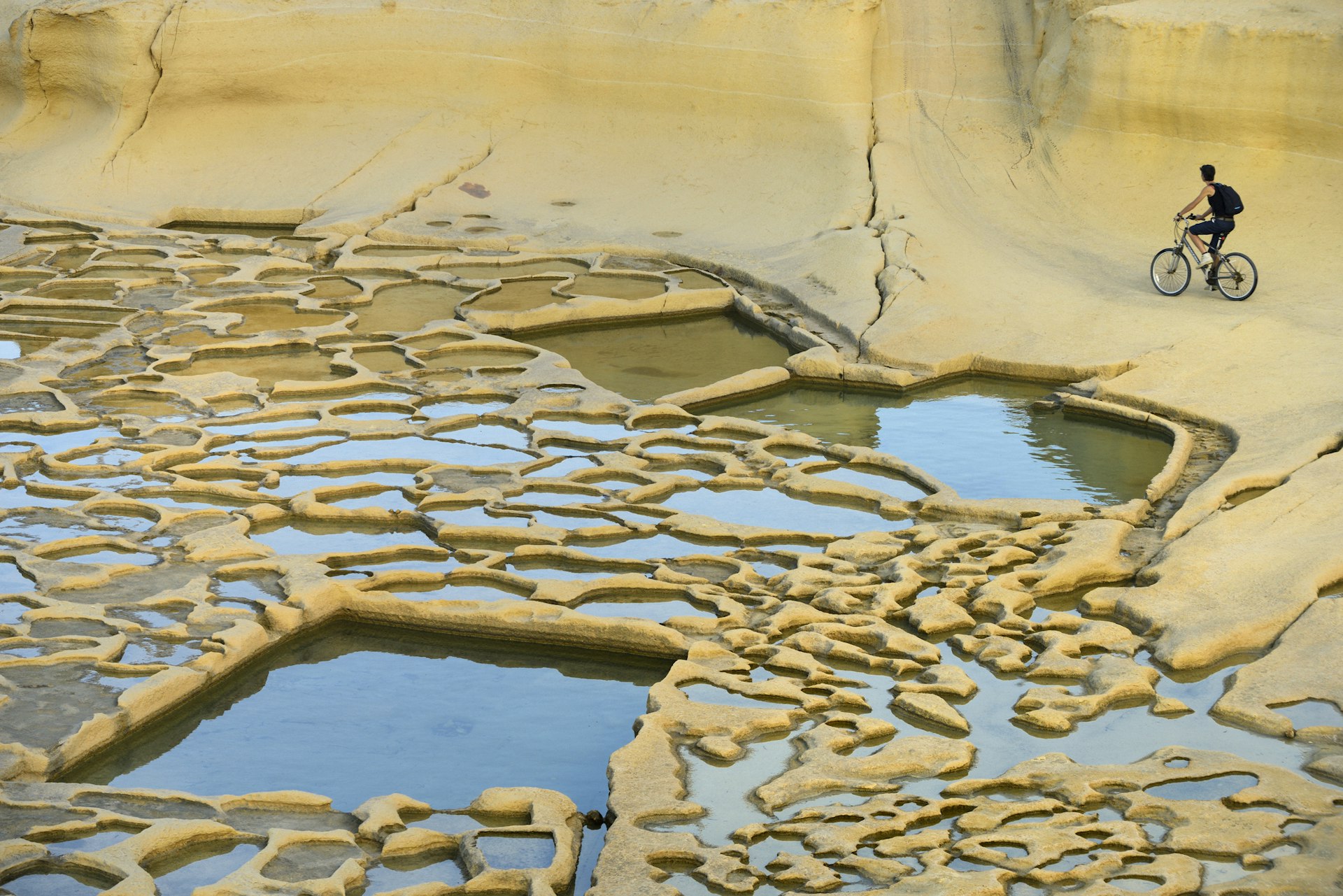 A solo cyclist pedals round a flat yellowish landscape in Gozo's salt pans