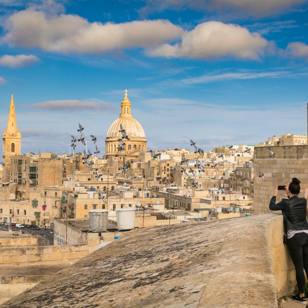 Two women tourists take photo of pigeons and beautiful skyline view of Capital city of Malta, Valletta, Dome and bell-tower above the walls, blue skies, from the St. Andrew Bastion, Valletta, Malta; Shutterstock ID 608541035; your: Claire Naylor; gl: 65050; netsuite: Online editorial; full: Malta best things to do