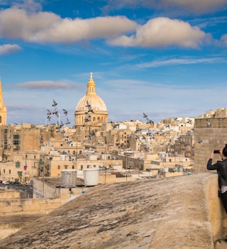 Two women tourists take photo of pigeons and beautiful skyline view of Capital city of Malta, Valletta, Dome and bell-tower above the walls, blue skies, from the St. Andrew Bastion, Valletta, Malta; Shutterstock ID 608541035; your: Claire Naylor; gl: 65050; netsuite: Online editorial; full: Malta best things to do