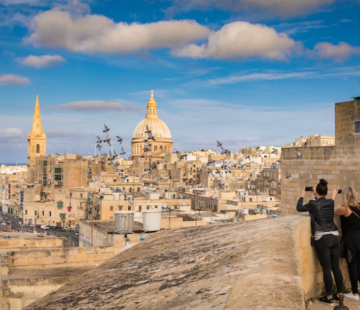Two women tourists take photo of pigeons and beautiful skyline view of Capital city of Malta, Valletta, Dome and bell-tower above the walls, blue skies, from the St. Andrew Bastion, Valletta, Malta; Shutterstock ID 608541035; your: Claire Naylor; gl: 65050; netsuite: Online editorial; full: Malta best things to do