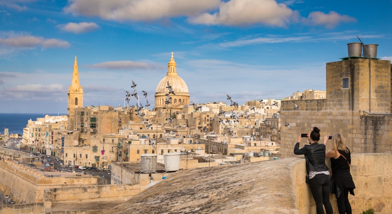 Two women tourists take photo of pigeons and beautiful skyline view of Capital city of Malta, Valletta, Dome and bell-tower above the walls, blue skies, from the St. Andrew Bastion, Valletta, Malta; Shutterstock ID 608541035; your: Claire Naylor; gl: 65050; netsuite: Online editorial; full: Malta best things to do