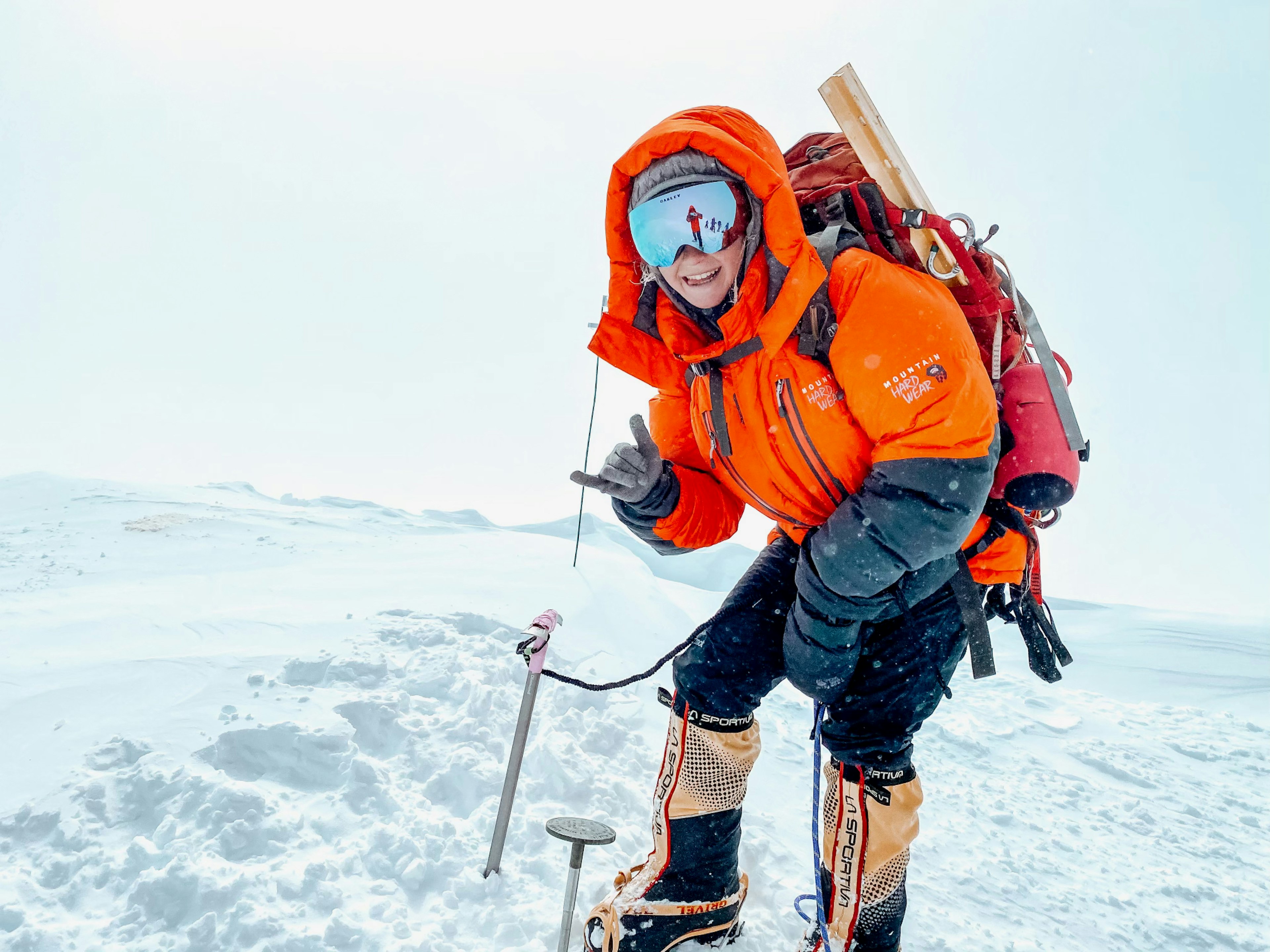 Lucy Westlake trekking through the snow in an orange and blue coat and reflective goggles, making the shaka sign with one hand