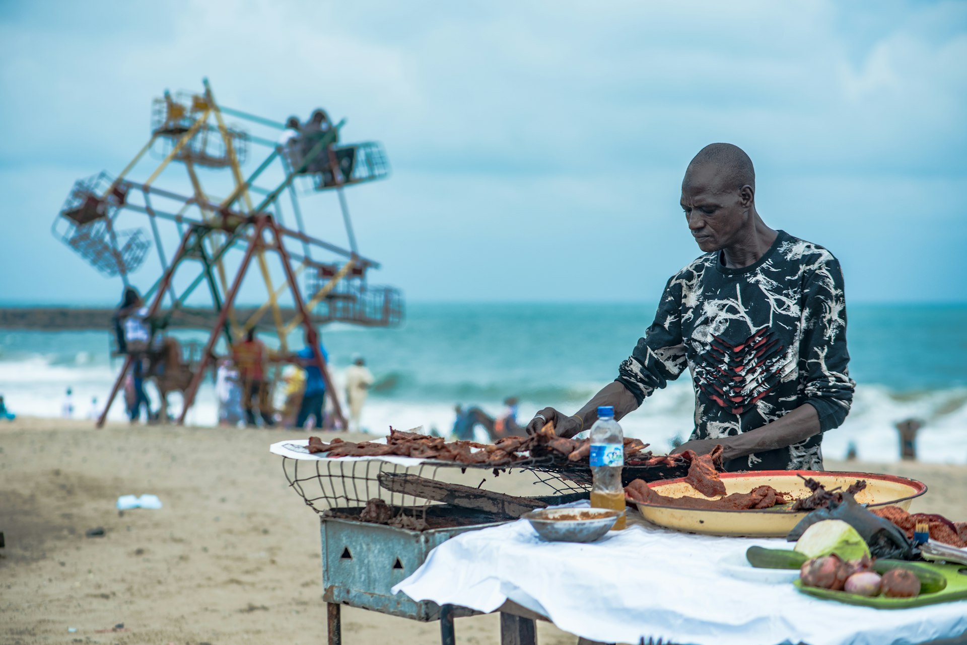 A chef barbecues at a grill by the Atlantic Ocean with a small Ferris wheel seen in the distance at Elegushi Beach, Lagos, Nigeria