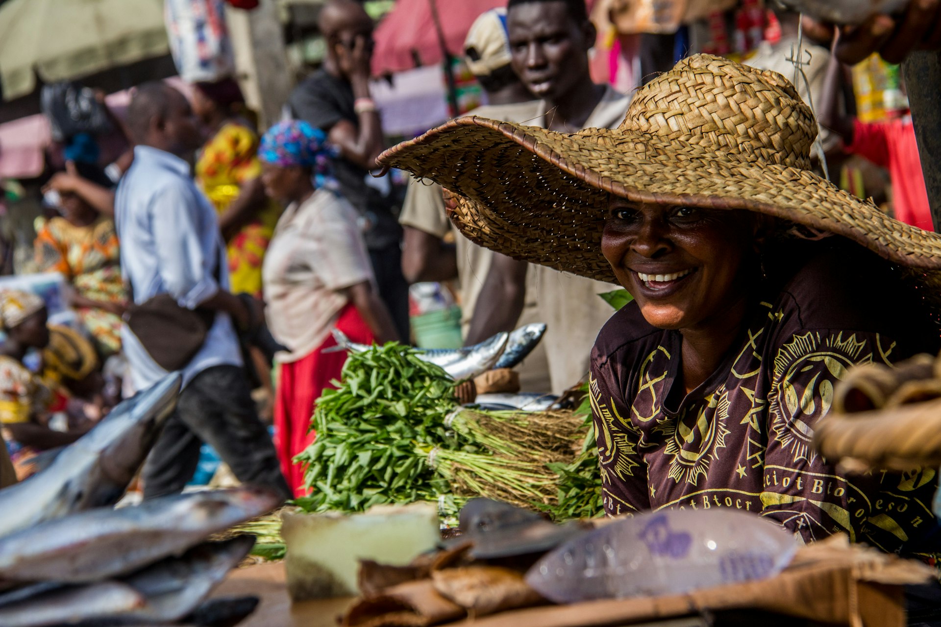 A vegetable vendor wearing a straw hat smiles at bustling Mile 12 Market in Lagos, Nigeria