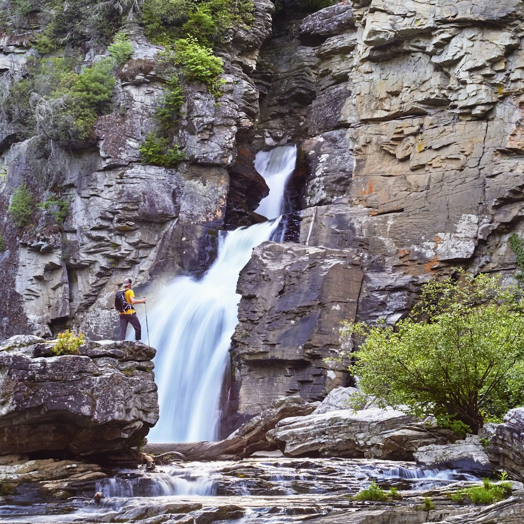 A hiker stands on a large rock in front of a waterfall, which is framed by rocky cliffs