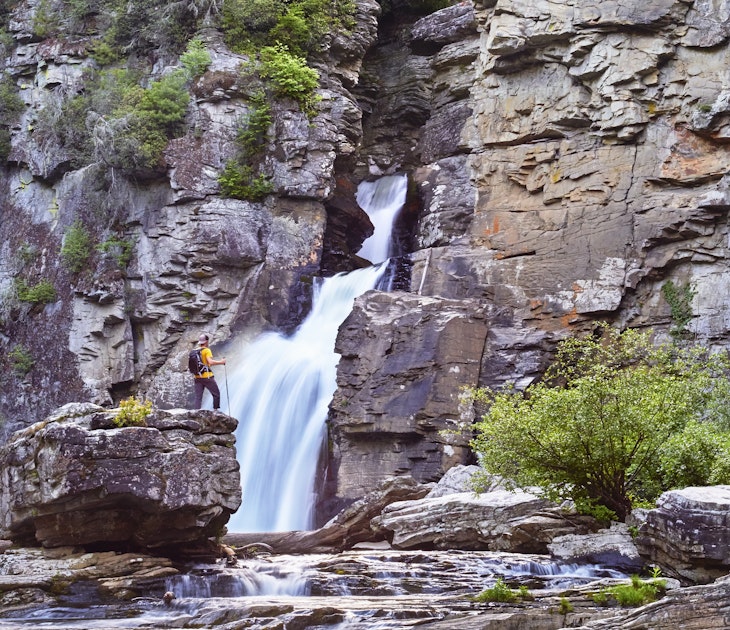 A hiker stands on a large rock in front of a waterfall, which is framed by rocky cliffs