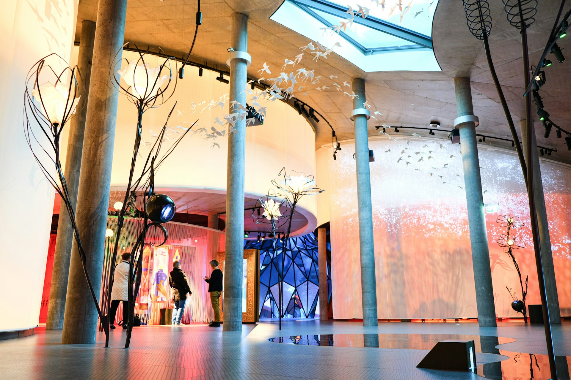 A large colourful curved wall with displays of light, with sculptures of giant flowers and bids soaring above