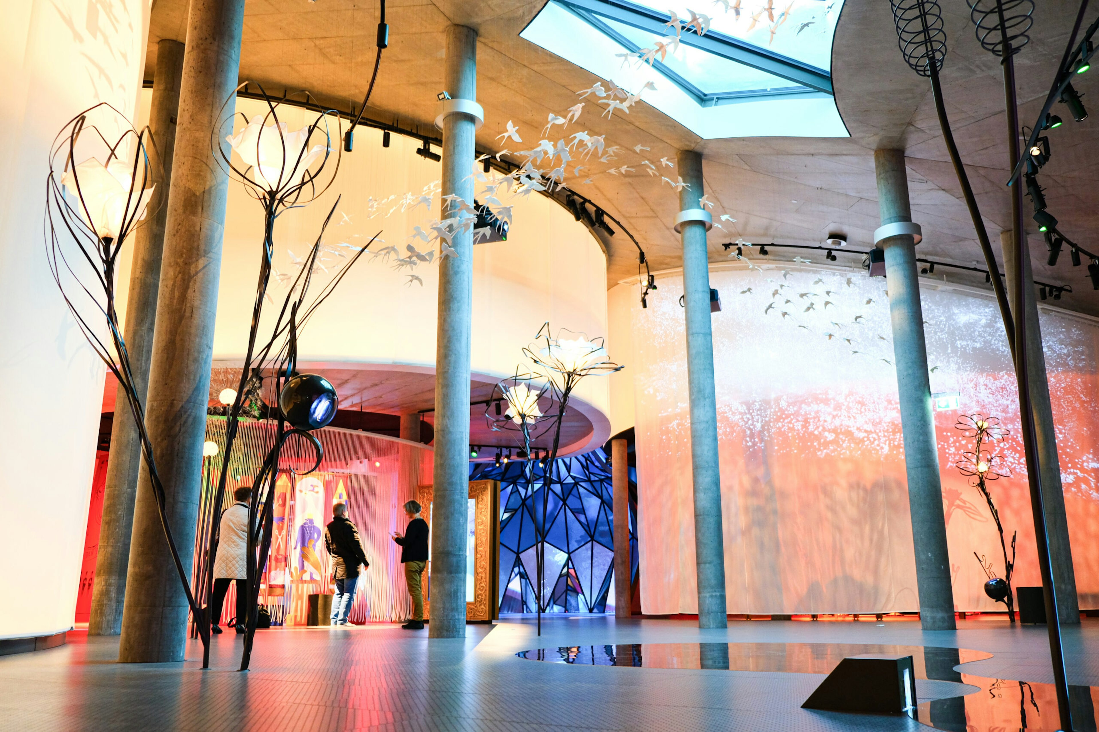 A large colourful curved wall with displays of light, with sculptures of giant flowers and birds soaring above