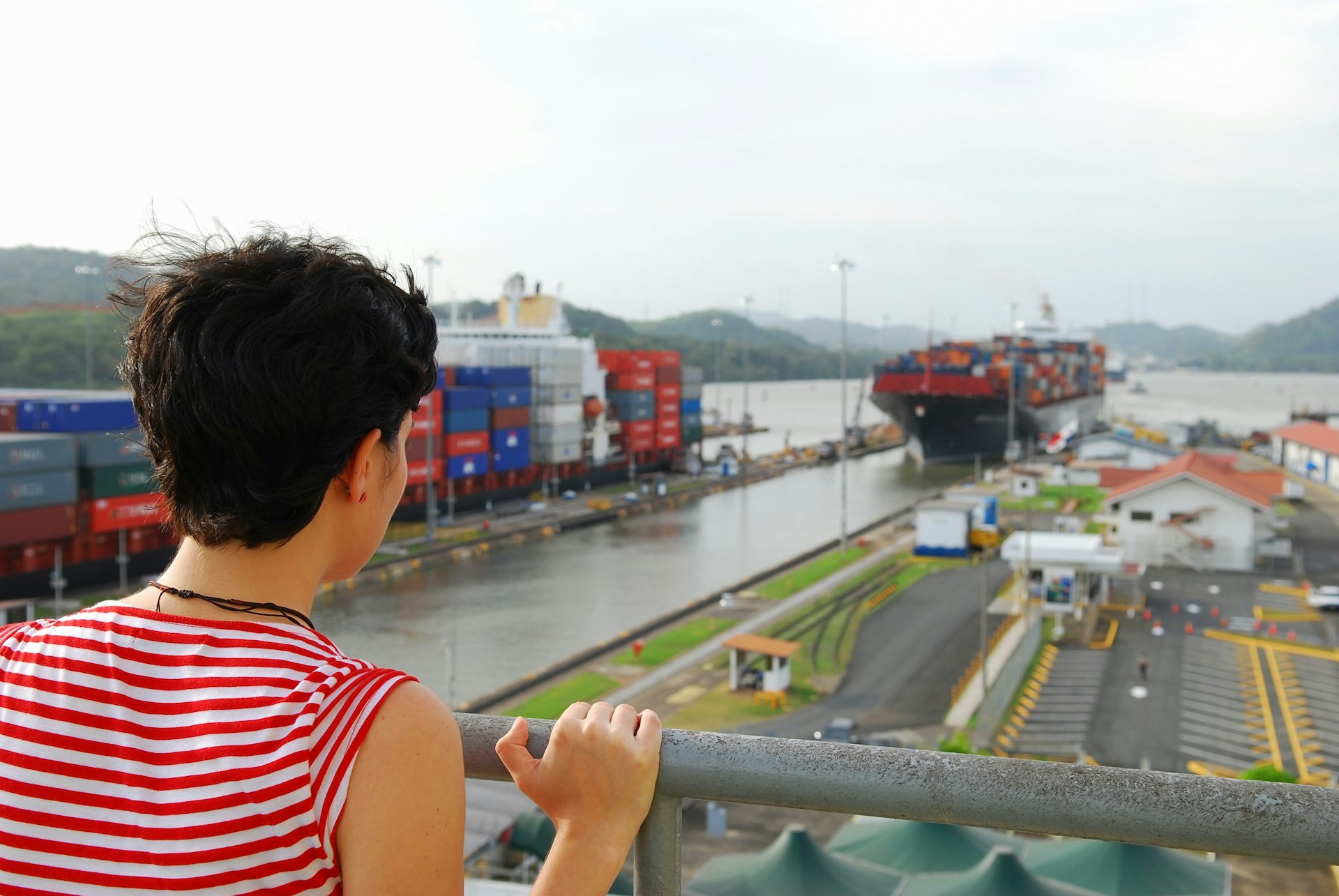 A young woman standing at the Miraflores Locks visitor viewing platform on the Panama Canal in Panama. She is watching two container ships entering the locks.