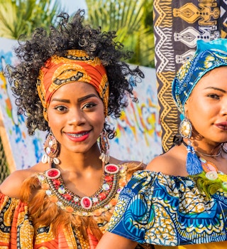 Two wearing colorful tradition garb and head scarves smile at the camera. 