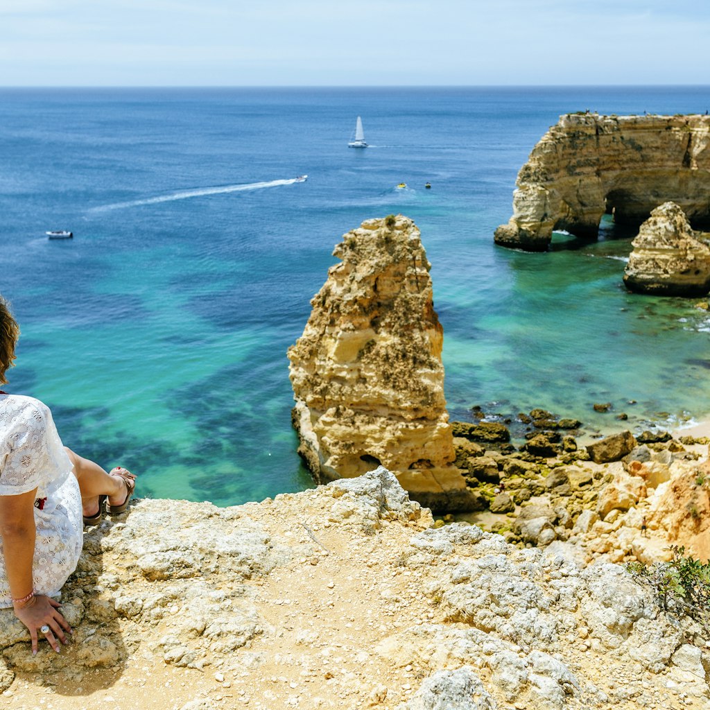 A woman sits on a cliff looking towards at the jagged cliff line and rock formations of of Marina beach in the Algarve, Portugal