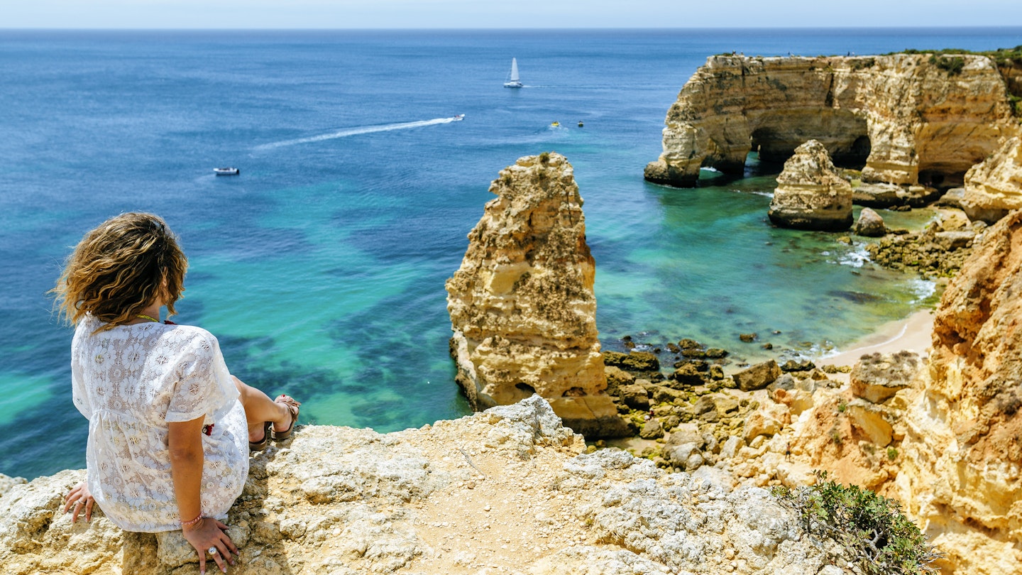 A woman sits on a cliff looking towards at the jagged cliff line and rock formations of of Marina beach in the Algarve, Portugal