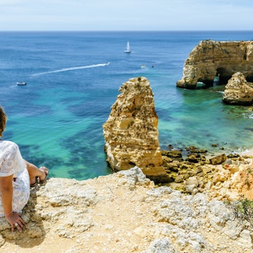 A woman sits on a cliff looking towards at the jagged cliff line and rock formations of of Marina beach in the Algarve, Portugal