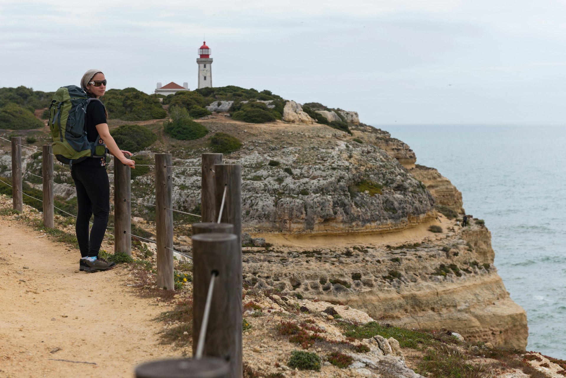 A female hiker on the Seven Hanging Valleys Trail (Percurso dos Sete Vales Suspensos) admires a view of a lighthouse and striated rock formations, the Algarve, Portugal