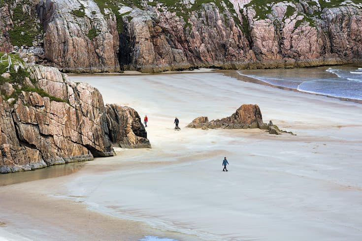 Walkers in a sandy bay with huge rocks towering over them, Rispond Bay, Durness, Scotland