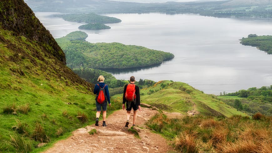Two hikers head down a hill towards a large body of water, Loch Lomond in Scotland
