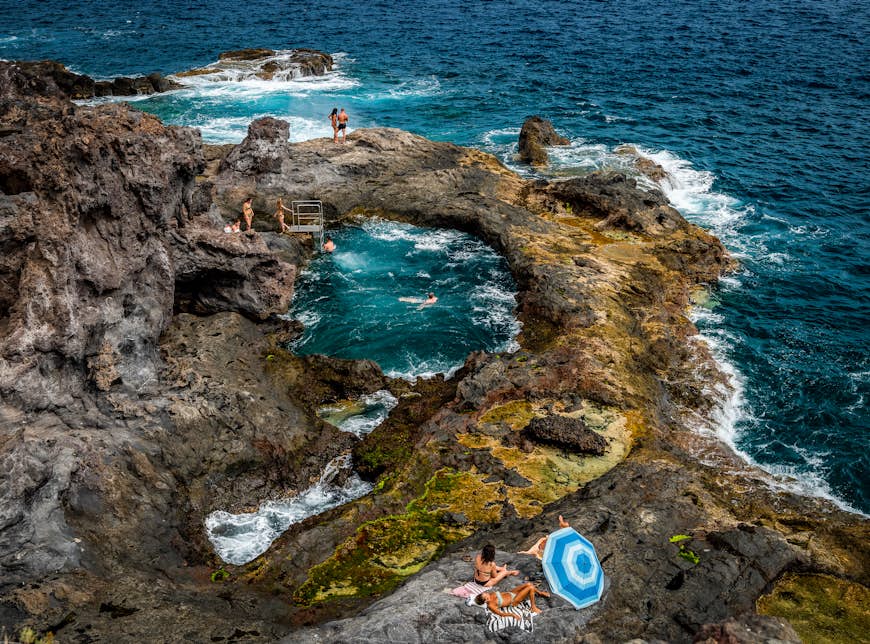 Tourists and locals enjoy swimming and sunbathing in a natural saltwater pool, formed in an area of volcanic cliffs
