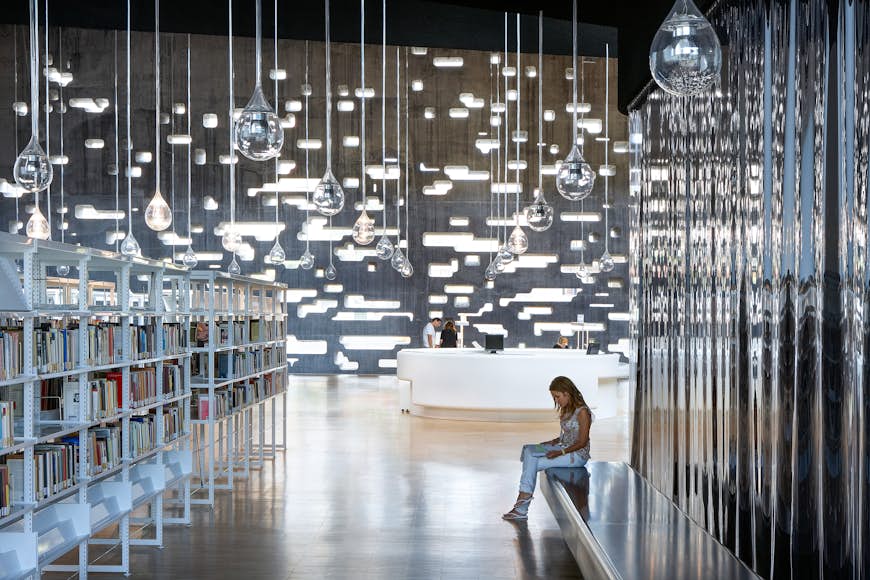 A woman sits in the library at the Tenerife Espacio De Las Artes. Glass sculptures that look like raindrops hang from the ceiling and a modern white desk is visible in the background. 