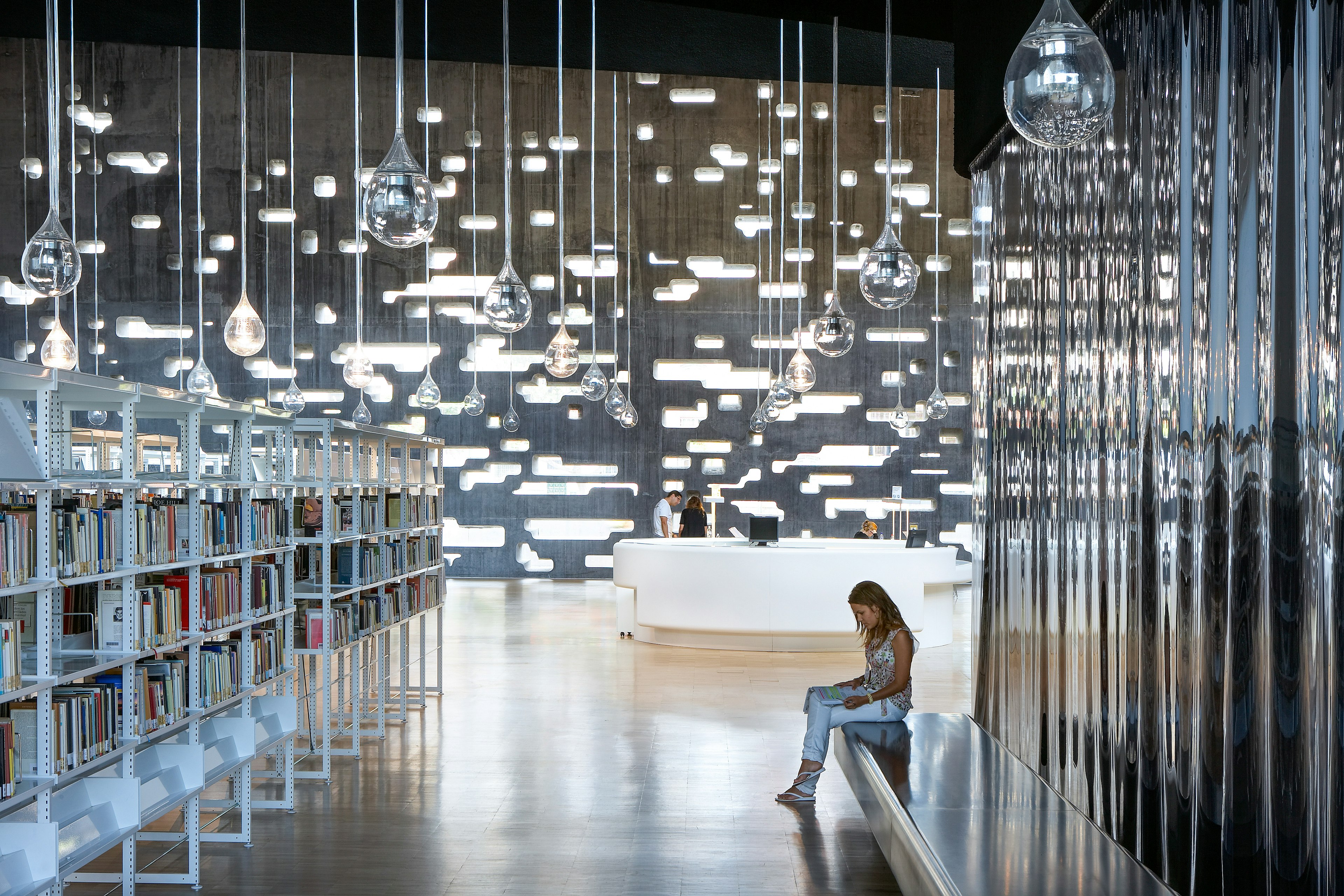 A woman sits in the library at the Tenerife Espacio De Las Artes. Glass sculptures that look like raindrops hang from the ceiling and a modern white desk is visible in the background.