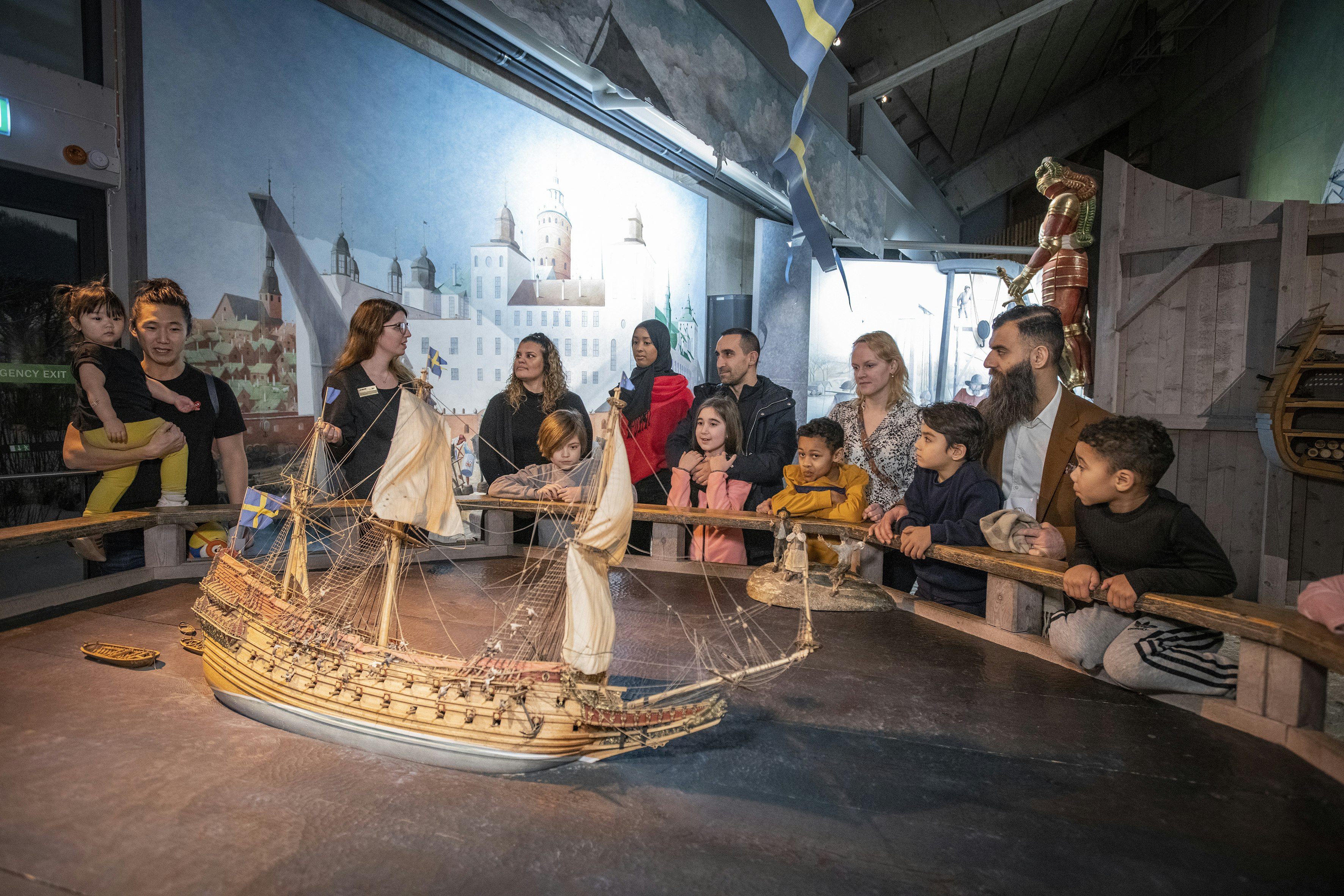 A multi-ethnic group of tourists gather round a model of a ship and listen to a museum guide at Stockholm's Vasa Museum