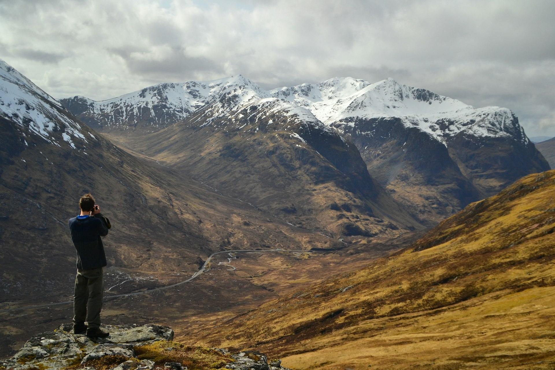 A man takes a photo of a vista of snow-capped mountains on the West Highland Way, Scotland, United Kingdom