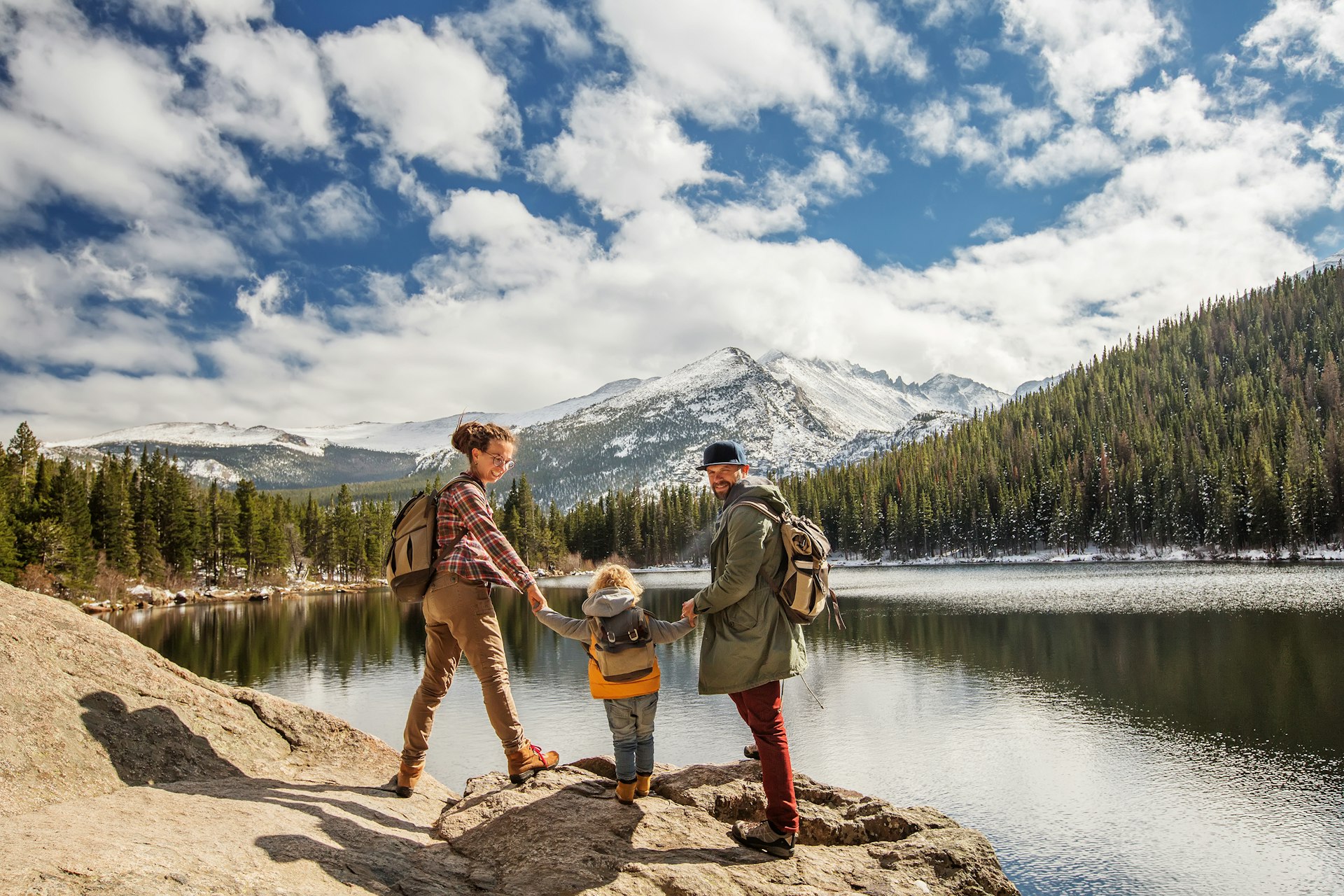 Two parents and a child hike by lake with pines and snowy mountains in the distance at Happy family in Rocky Mountain National Park, Colorado, USA 