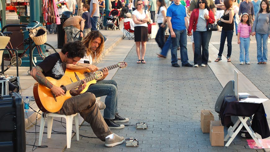 Two guitarists busk in a pedestrianized zone with plenty of onlookers