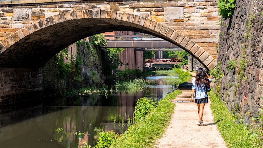 The back of a woman as she follows a tow path alongside a canal approaching a stone bridge on the C & O Canal near Washington DC