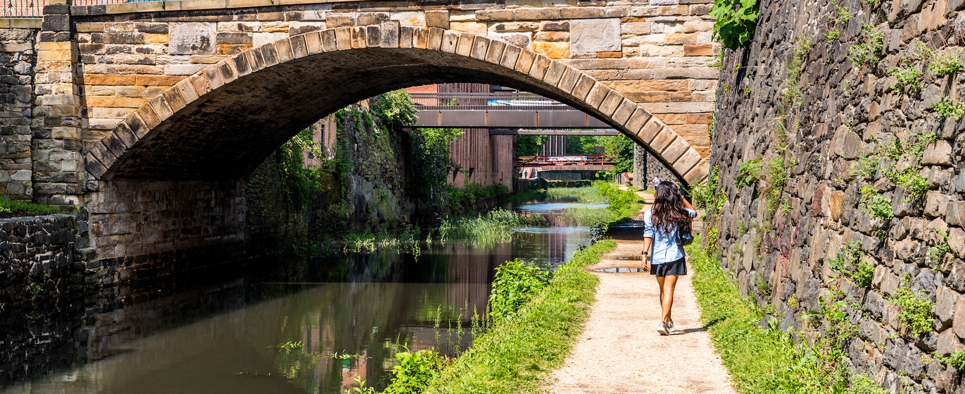 The back of a woman as she follows a tow path alongside a canal approaching a stone bridge on the C & O Canal near Washington DC