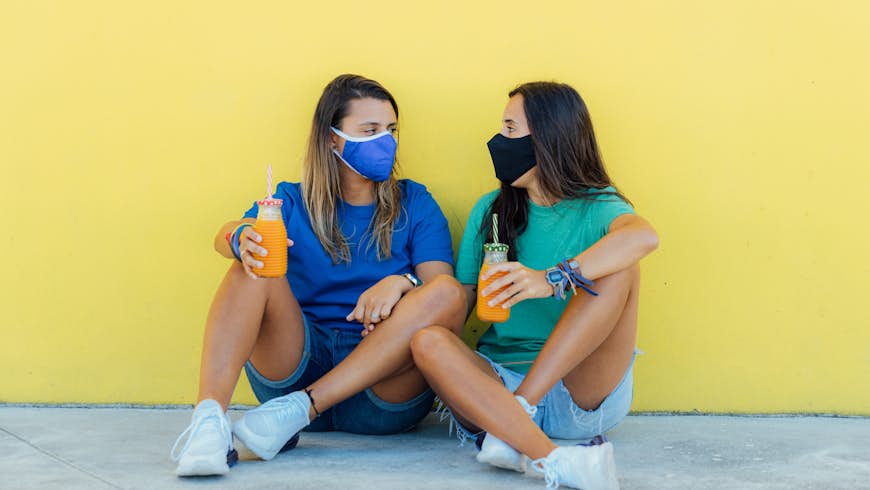 Young lesbian couple sitting wearing medical face masks, holding orange juices with a yellow wall in the background
