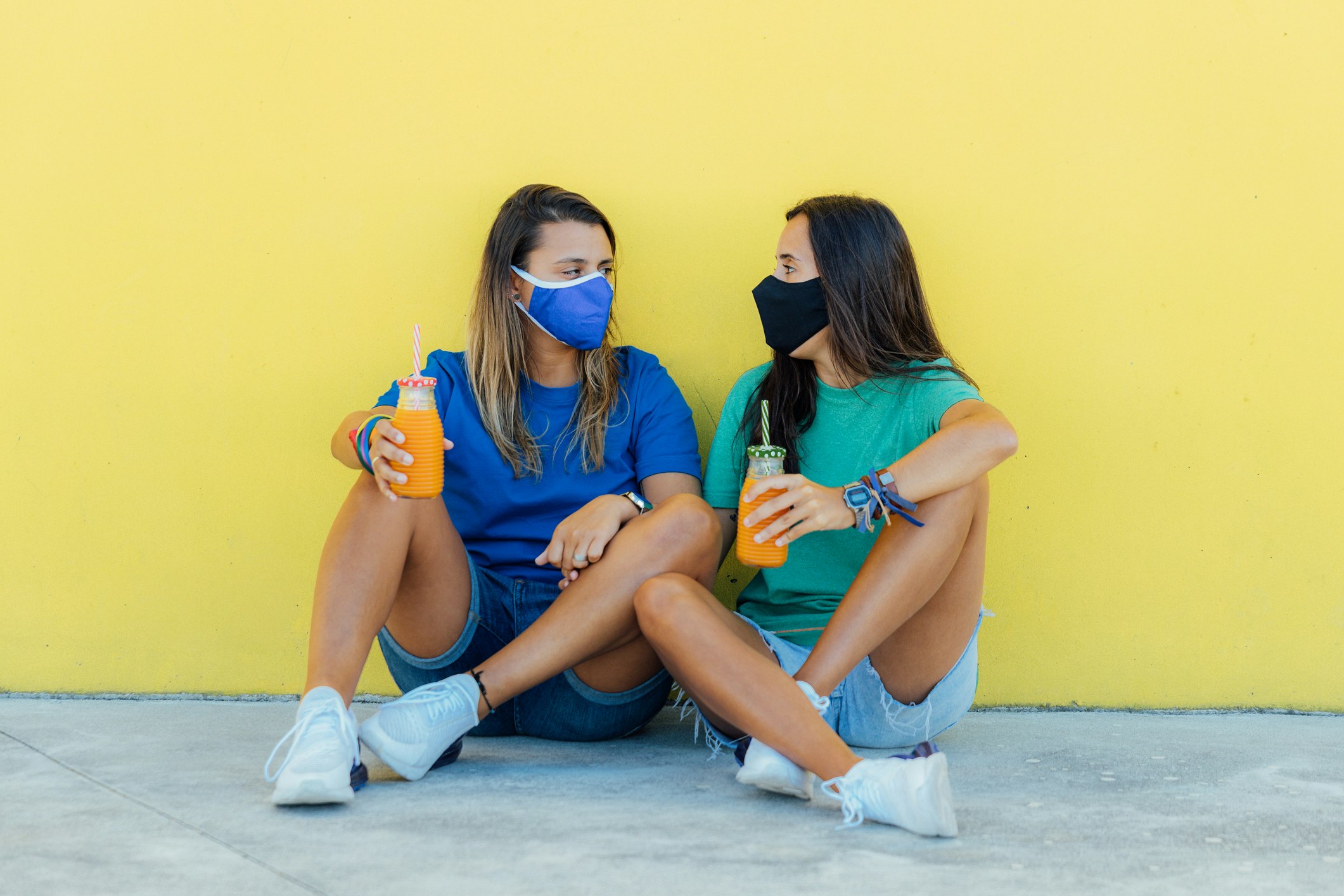 Young lesbian couple sitting wearing medical face masks, holding orange juices with a yellow wall in the background