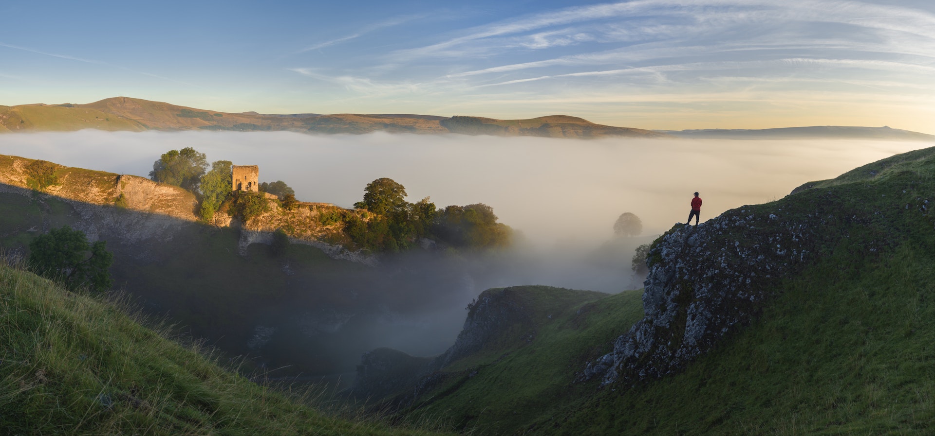 A lone figure stands on a rocky outcrop looking towards a ruined medieval castle on a foggy morning at the sun rises
