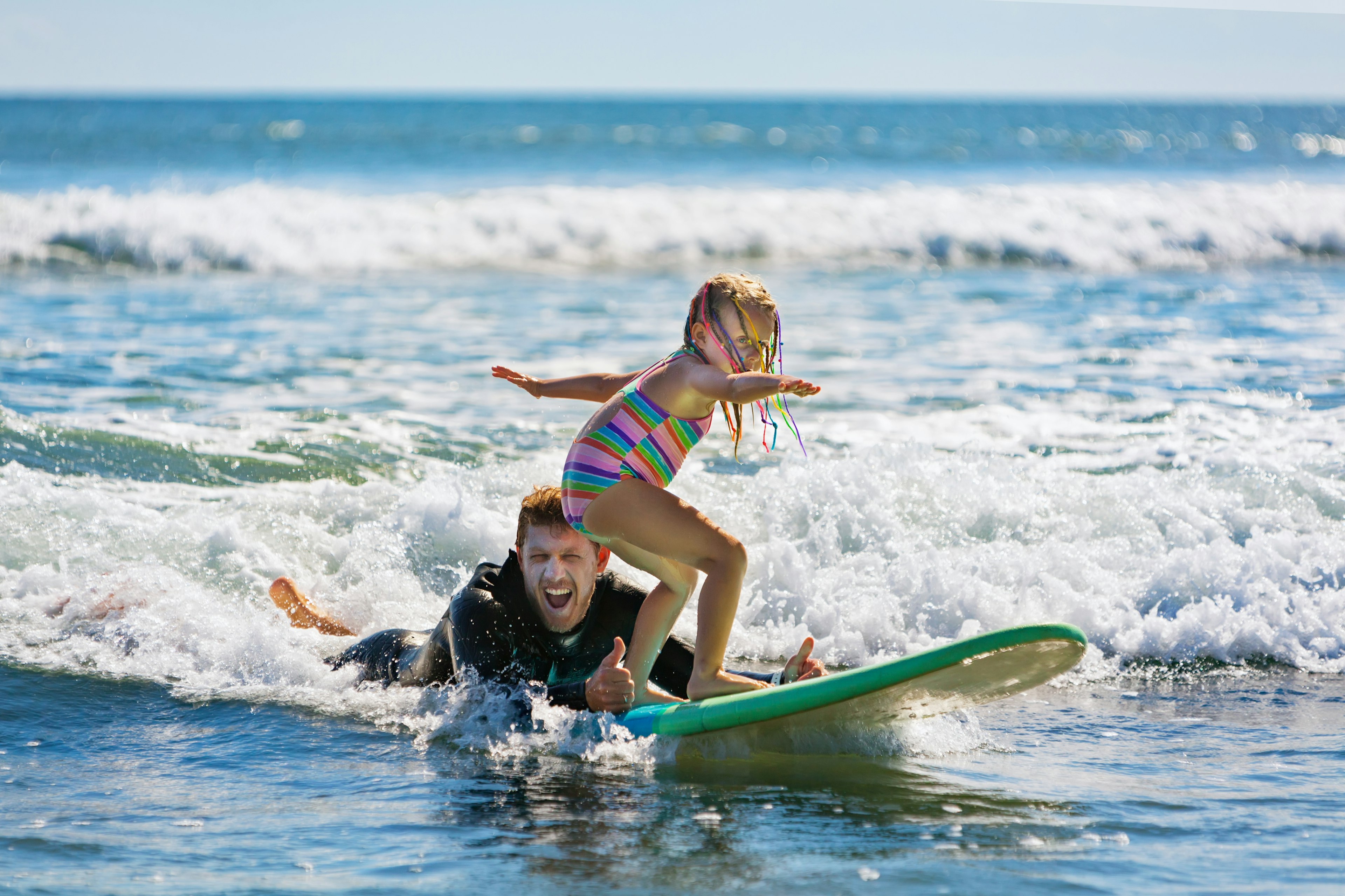 A man in a wet suit lies in the surf giving a thumbs-up signal as a young child balances on a surf board