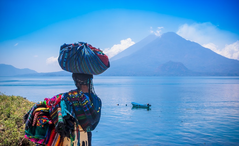 Panajachel, Guatemala -April, 25, 2018: Outdoor view of unidentifed indigenous woman, wearing typical clothes and walking in lakeshore with small boats in Atitlan Lake and volcano in Background in