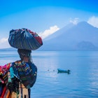 Panajachel, Guatemala -April, 25, 2018: Outdoor view of unidentifed indigenous woman, wearing typical clothes and walking in lakeshore with small boats in Atitlan Lake and volcano in Background in