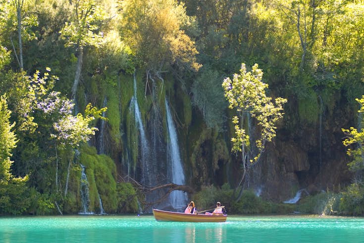 Two people sit in a small rowing boat in an incredibly turquoise lake with a waterfall