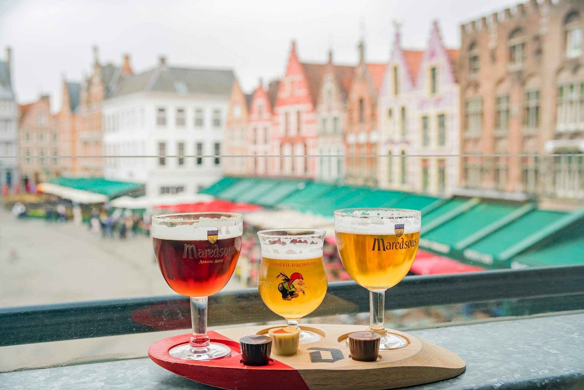 Three stemmed glasses of beer on a ledge overlooking a Belgian street with colorful houses