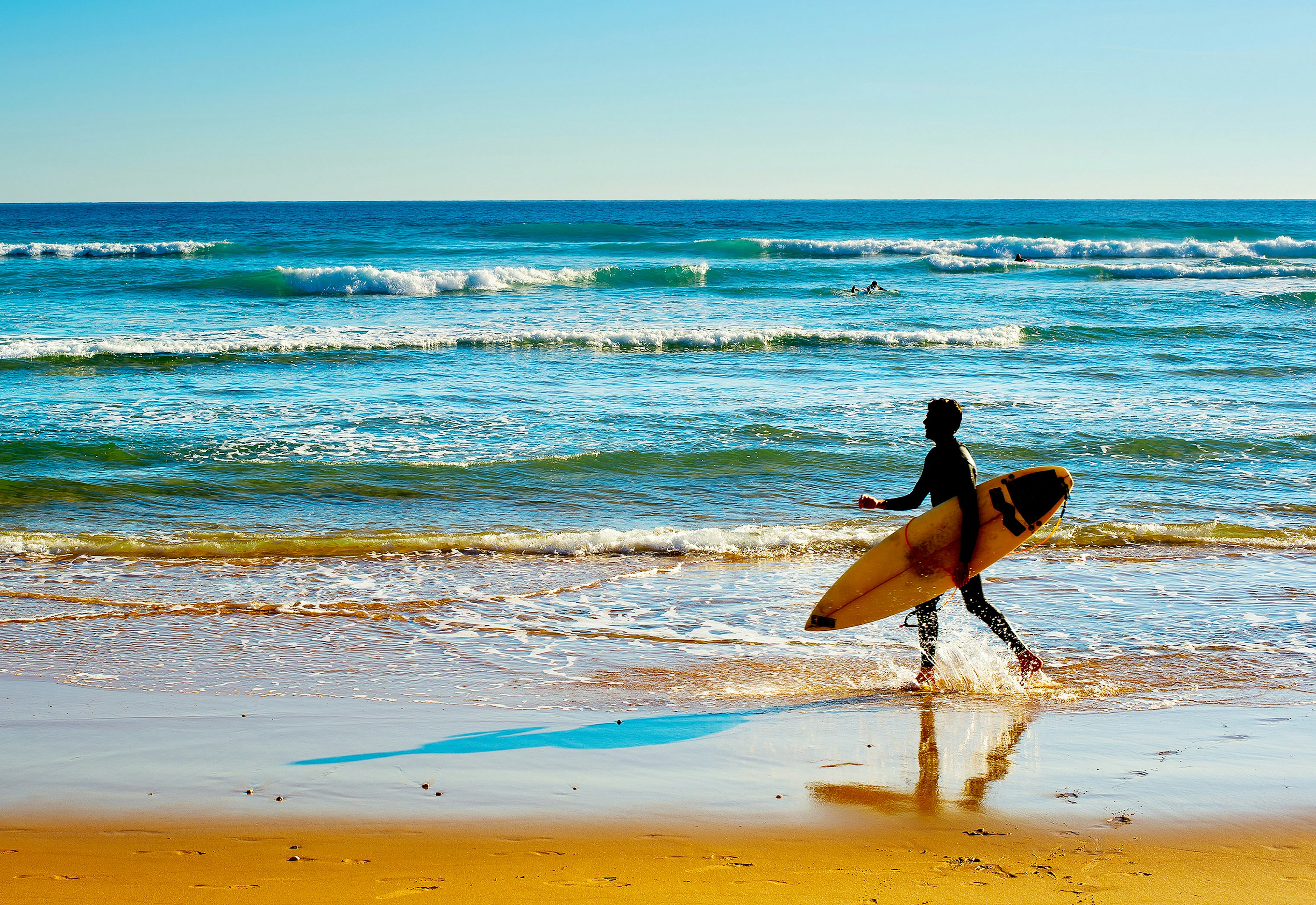 A surfer in silhouette on the beach carrying a surf board as the waves crash behind them