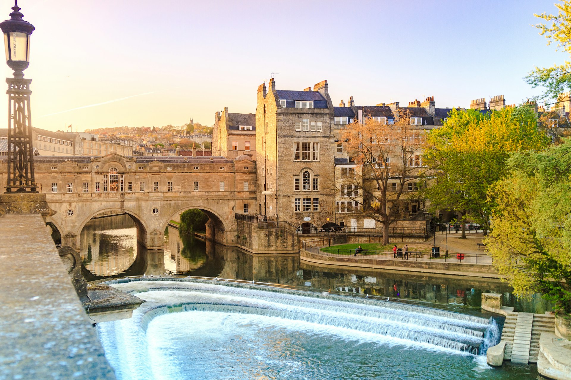 The stone Pulteney Bridge spans over the River Avon as the sun sets in the background. The bridge has curved arches underneath and windows along the side where the shops are.