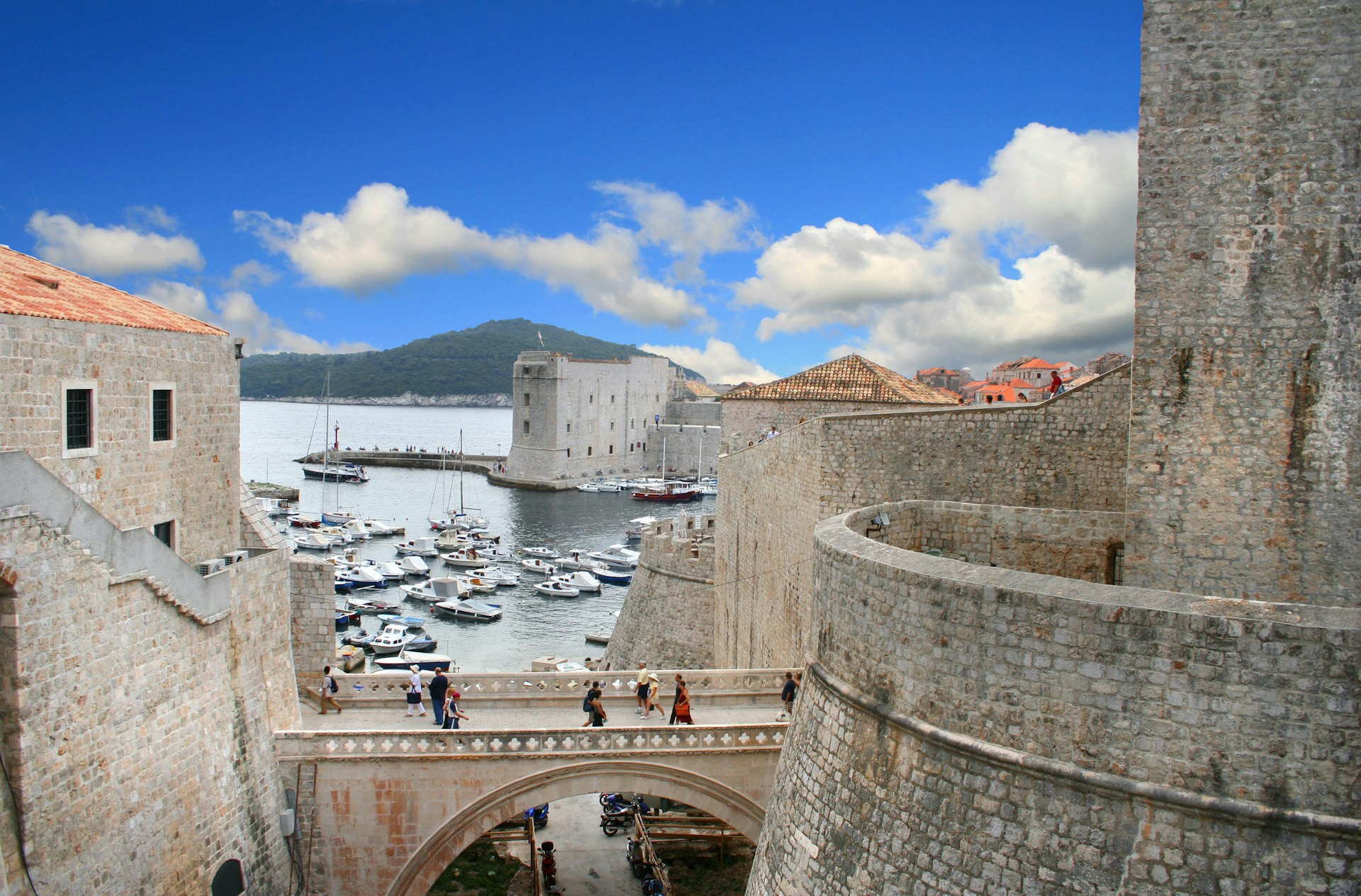 Gente cruzando un puente de piedra en Dubrovnik con el puerto más allá