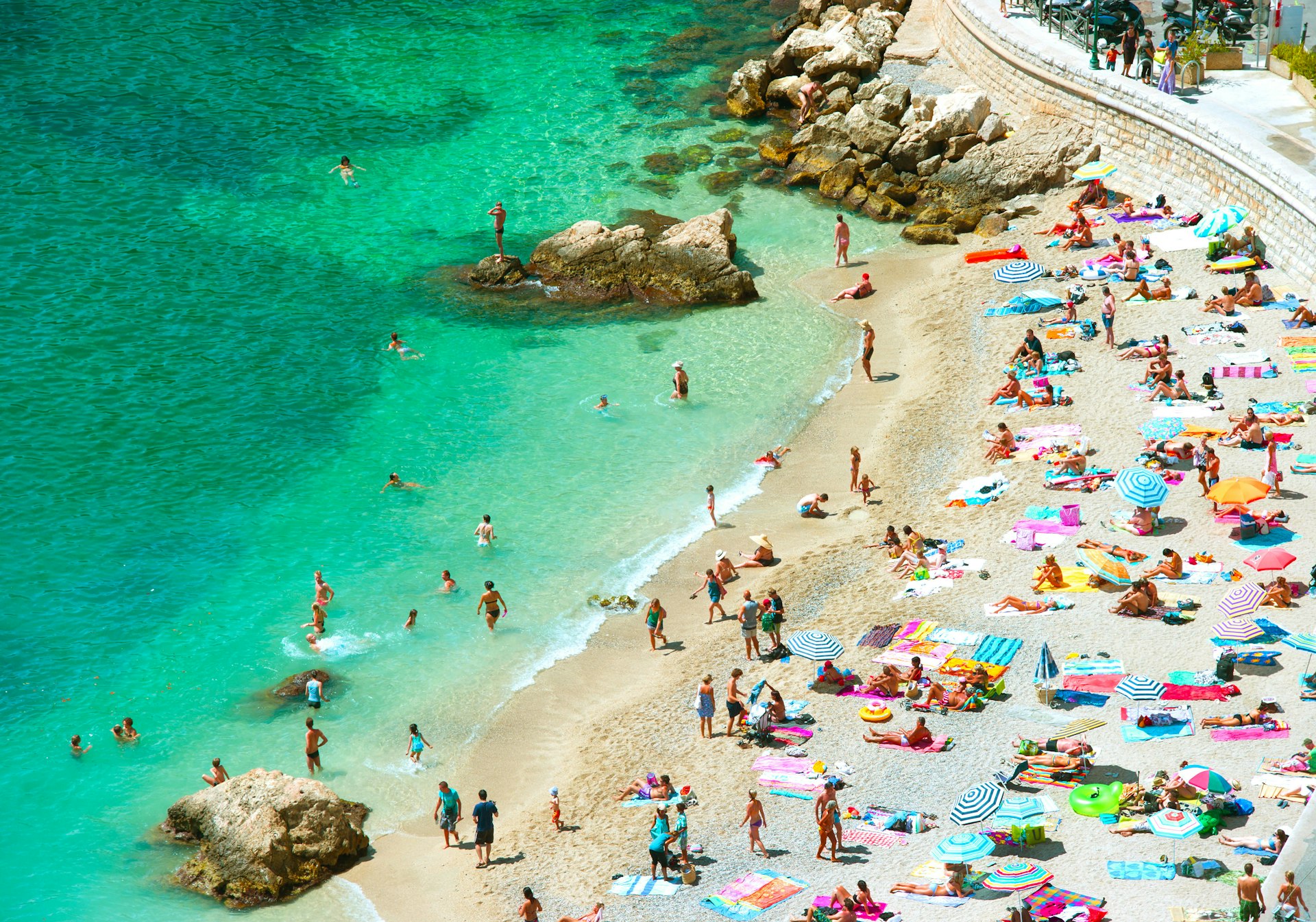 An aerial shot of the crowded beach and water with many tourists and brightly colored sun umbrellas at Villefranche-sur-Mer, Côte d’Azur, France, Europe