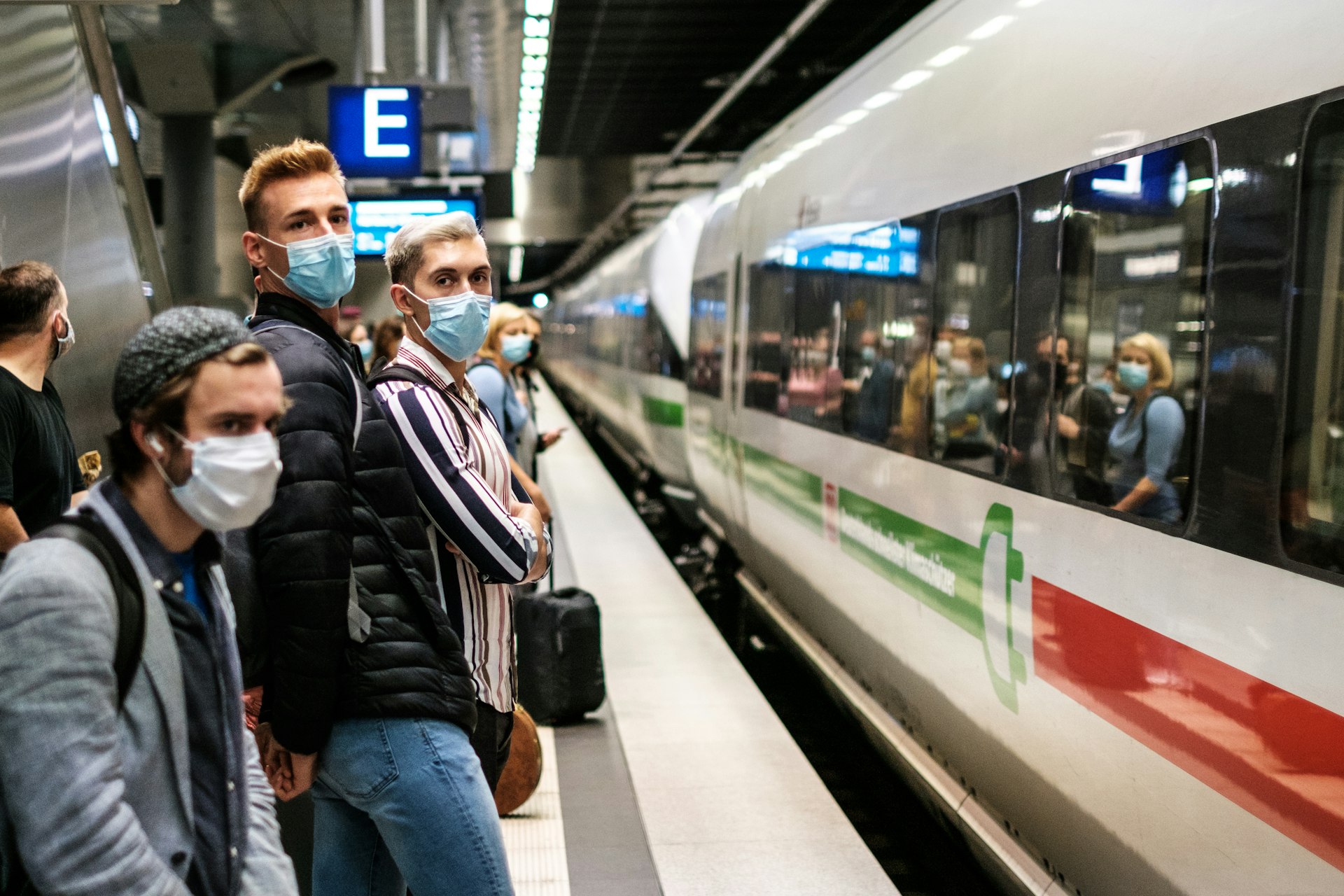 People stand on the platform wearing masks and waiting for a train. 