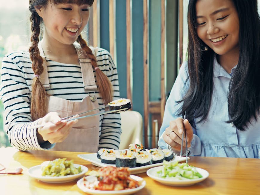 Two Asian women eating gimbap (Korean sushi) with plates of kimchi