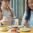 Asian women holding silver chopsticks to eat Korean seaweed rice roll with cheese or cheese kimbap (sometimes spelled gimbap) and kimchi on wooden table.